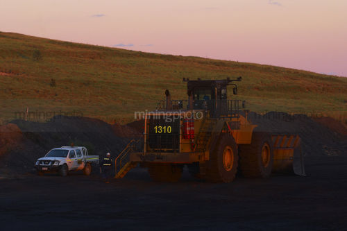Mine site worker walking around parked loader at the end of a shift.  light vehicle parked adjacent. shot in late afternoon dusk light. - Mining Photo Stock Library