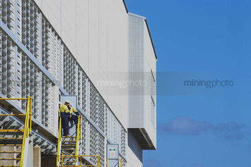 Two mine site workers in discussion standing on a  platform at the edge of shed platform.  blue sky behind. - Mining Photo Stock Library