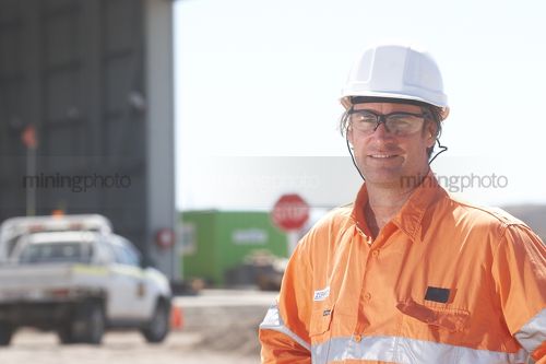 Mine site worker in full PPE looking straight at the camera in front of workshop. light vehicle driving in background.  space for copy on left hand side. - Mining Photo Stock Library
