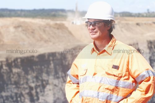 Mine site worker in full PPE standing with hands on hips at open cut mining site.  high wall of coal in background.  worker's face clearly visible. - Mining Photo Stock Library