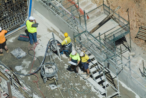 Aerial of workers in PPE on dam wall construction site looking at plans and giving radio instructions.  scaffolding and work concrete tools adjacent. - Mining Photo Stock Library