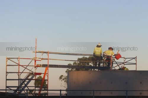 Workers in full PPE heading into confined space from scaffolding.  shot at dawn and from a distance away. - Mining Photo Stock Library