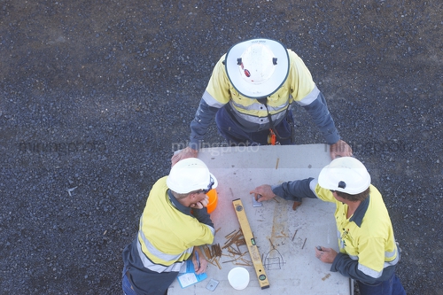 Construction safety meeting.  aerial photo of 3 workers in full PPE including hard hats and long sleeves having a safety meeting. could be on a construction mine site.  faces cannot be seen.  workers are drawing out designs. - Mining Photo Stock Library