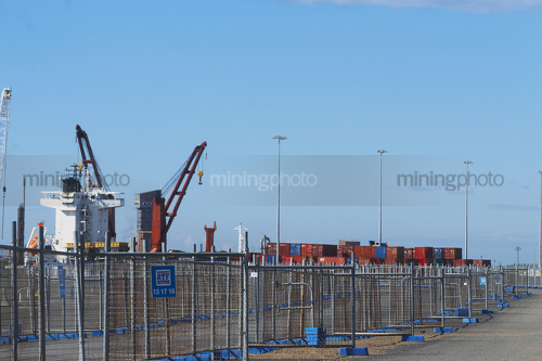 Photo of temporary fencing around ship at port.  shipping containers stacked on wharf.  bluer sky behind. - Mining Photo Stock Library