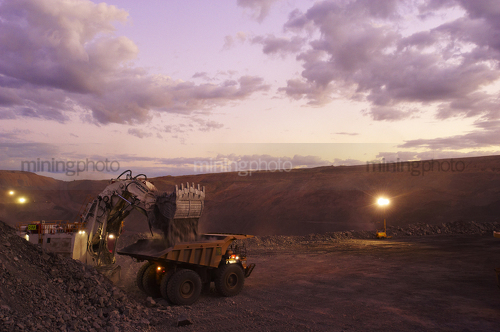 Great dusk photo of a digger loading a haul truck at open cut mine site.  lights are on. - Mining Photo Stock Library