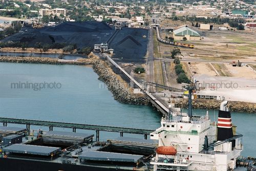 Coal being loaded by conveyor onto ship with heavy rail train and coal carriages moving in background. - Mining Photo Stock Library