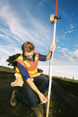 Road surveyor in ppe checking levels of rural road during upgrade. blue sky - Mining Photo Stock Library