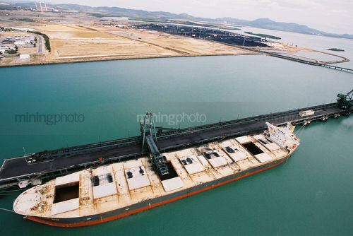 Coal ship being loaded out at a wharf by ship loader with coal stockpile and industry on land in the beackground - Mining Photo Stock Library