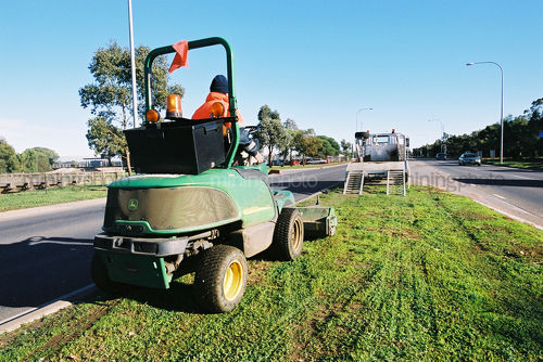 Maintenance worker in PPE on small tractor mowing centre grasas strip on residential highway road. - Mining Photo Stock Library