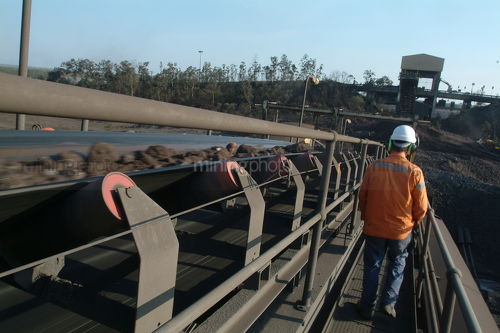 Worker at mine site walking next to conveyor with product. shot from behind - Mining Photo Stock Library