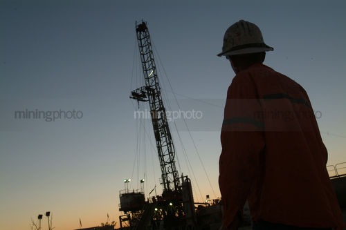 Silhouette of a drill rig worker with the derrick behind at dusk. - Mining Photo Stock Library