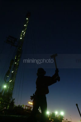 Silhouette of drill rig worker hammering with the lights of the  derrick behind at dusk. - Mining Photo Stock Library