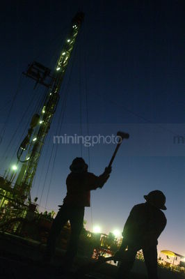 Silhouette of 2 drill rig workers hammering with the lights of the  derrick behind at dusk. - Mining Photo Stock Library