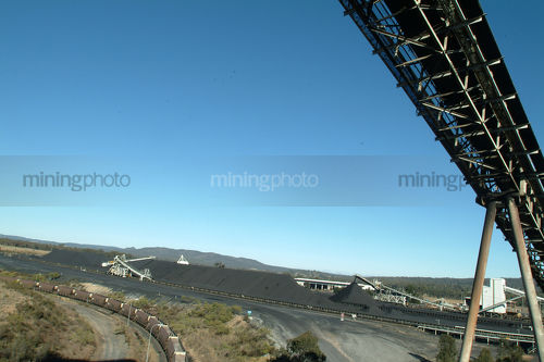 Coal conveyor loader above with heavy rail train carriage below and coal stockpiles in background. - Mining Photo Stock Library