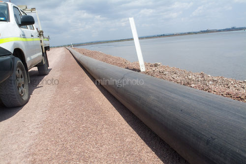 Light vehicle on road next to water pipe and large dam with survey peg. - Mining Photo Stock Library