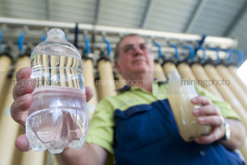 Man in PPE holding plastic bottle full of clean water at treatment plant with water purification process in background. - Mining Photo Stock Library