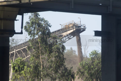 Moving conveyor stockpiling product at plant with some vegetation and trees in foreground - Mining Photo Stock Library