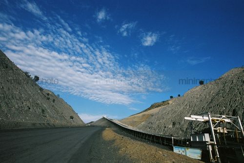 Conveyor leaving underground mine and traveling up graded road.  blue sky in background.  - Mining Photo Stock Library