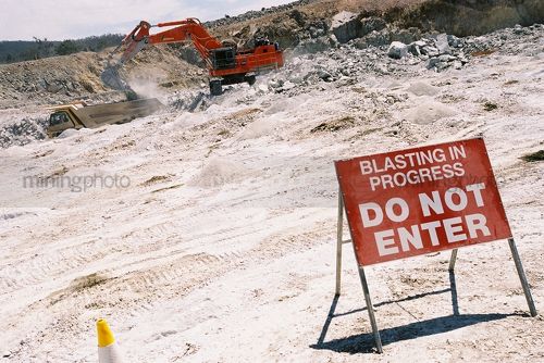 Excavator loading haul trucks next to signed blasting area. - Mining Photo Stock Library