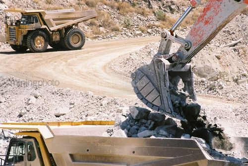 Excavator bucket loading hard metal into haul truck with empty truck in background on haul road. - Mining Photo Stock Library