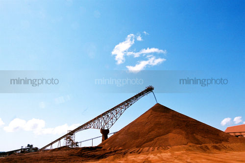 Conveyor stockpiling red dirt with brilliant blue sky behind. - Mining Photo Stock Library