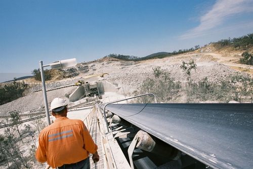 Worker walking down the conveyor walkway with open cut mine site in background.  shot from behind. - Mining Photo Stock Library