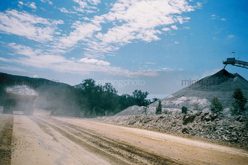 Loaded haul truck leaving cloud of dust as it drives along road with conveyor stockpiling in background.  great shot.  - Mining Photo Stock Library