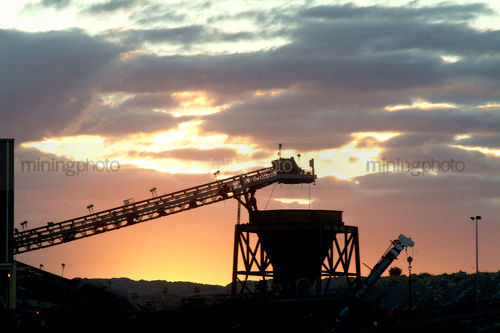 Silhouette of a conveyor delivering product to hopper with sunset behind. - Mining Photo Stock Library