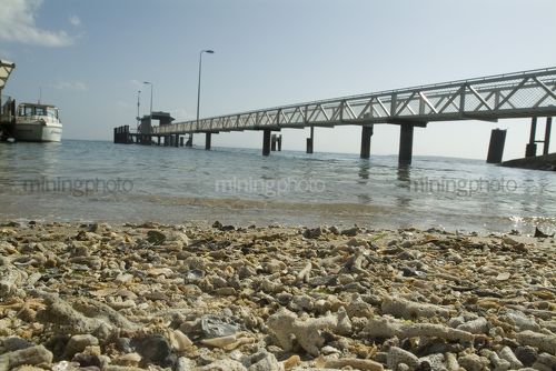 Pedestrian jetty out to passenger ferry terminal over ocean.  shot from water level with shells on the beach in the foreground. - Mining Photo Stock Library