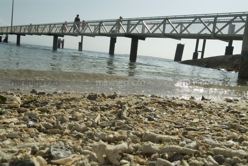 Person walking along pedestrian jetty out to passenger ferry terminal over ocean.  shot from water level with shells on the beach in the foreground. - Mining Photo Stock Library