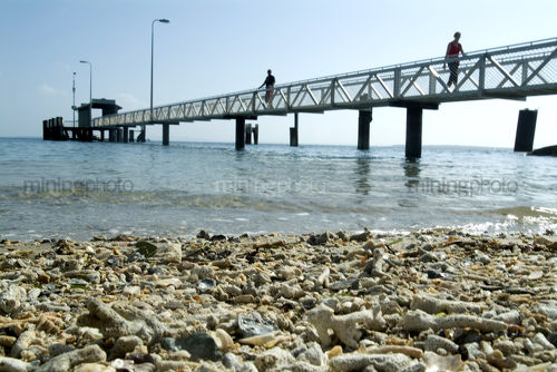 People walking along pedestrian jetty out to passenger ferry terminal over ocean.  shot from water level with shells on the beach in the foreground. - Mining Photo Stock Library