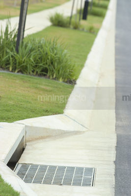 Closeup of storm water drain on the edge of a road with footpath and nature strip in background - Mining Photo Stock Library