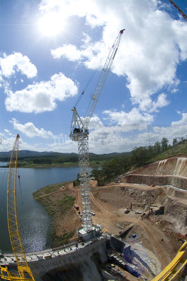Infrastructure dam building construction site with crane, earthworks, building with dam water behind. aerial vertical shot - Mining Photo Stock Library