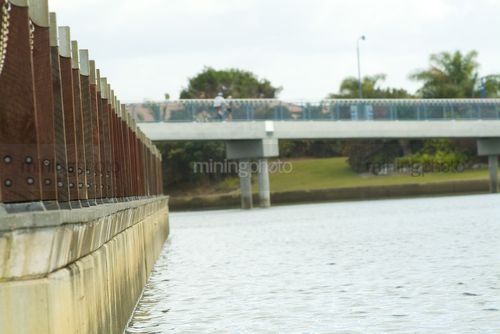 Edge of waterfront with pedestrian bridge in background. out of focus family riding and walking dogs in background. - Mining Photo Stock Library