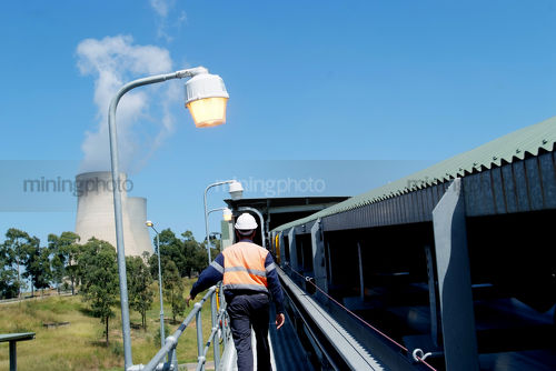 Mine worker on walkway next to covered coal conveyor with cooling towers from power station in background. shot from behind. - Mining Photo Stock Library