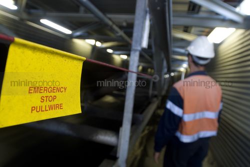 Worker in PPE walking next to covered conveyor with safety warning sign in foreground.  - Mining Photo Stock Library