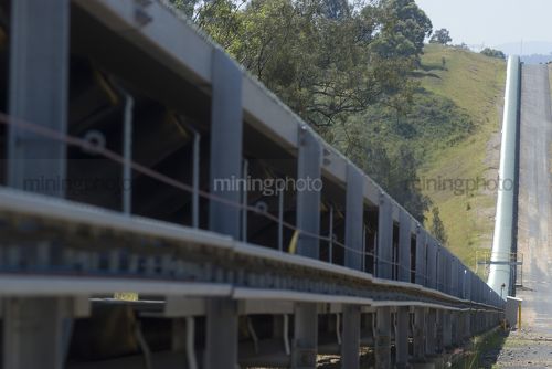 Covered, overland, coal, conveyor stretching for a long distance over hills.  sealed vehicle road access adjacent. - Mining Photo Stock Library
