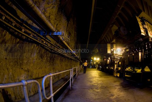 Worker underground walking away from camera with coal conveyors nearby - Mining Photo Stock Library