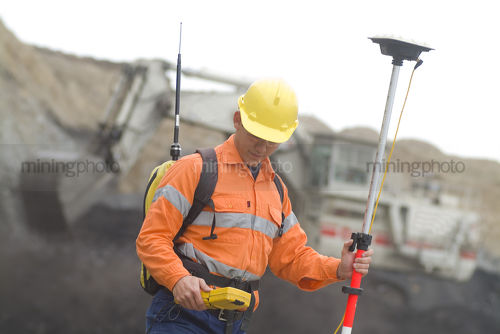 Close up of mine surveyor in full PPE with gear on open cut coal mine floor with excavator operating in background. vertical shot. - Mining Photo Stock Library