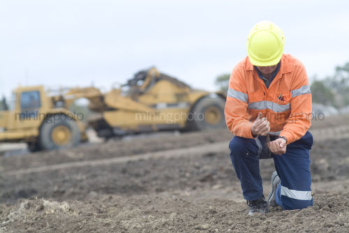 Mine worker on haunches sifting soil overburden through hands with scraper machinery in background. great generic mine shot with room for text. - Mining Photo Stock Library