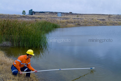Mine environment worker taking water samples from dam at open  cut mine.  haul truck on road in background. - Mining Photo Stock Library