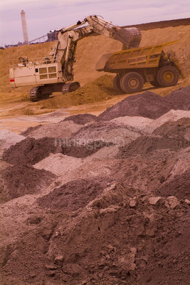 Excavator loading overburden into haul truck with coal fired power station smokestack in background. vertical shot. - Mining Photo Stock Library