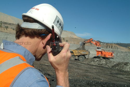 Mine site supervisor listening to radio with excavator loading haul truck in background. - Mining Photo Stock Library
